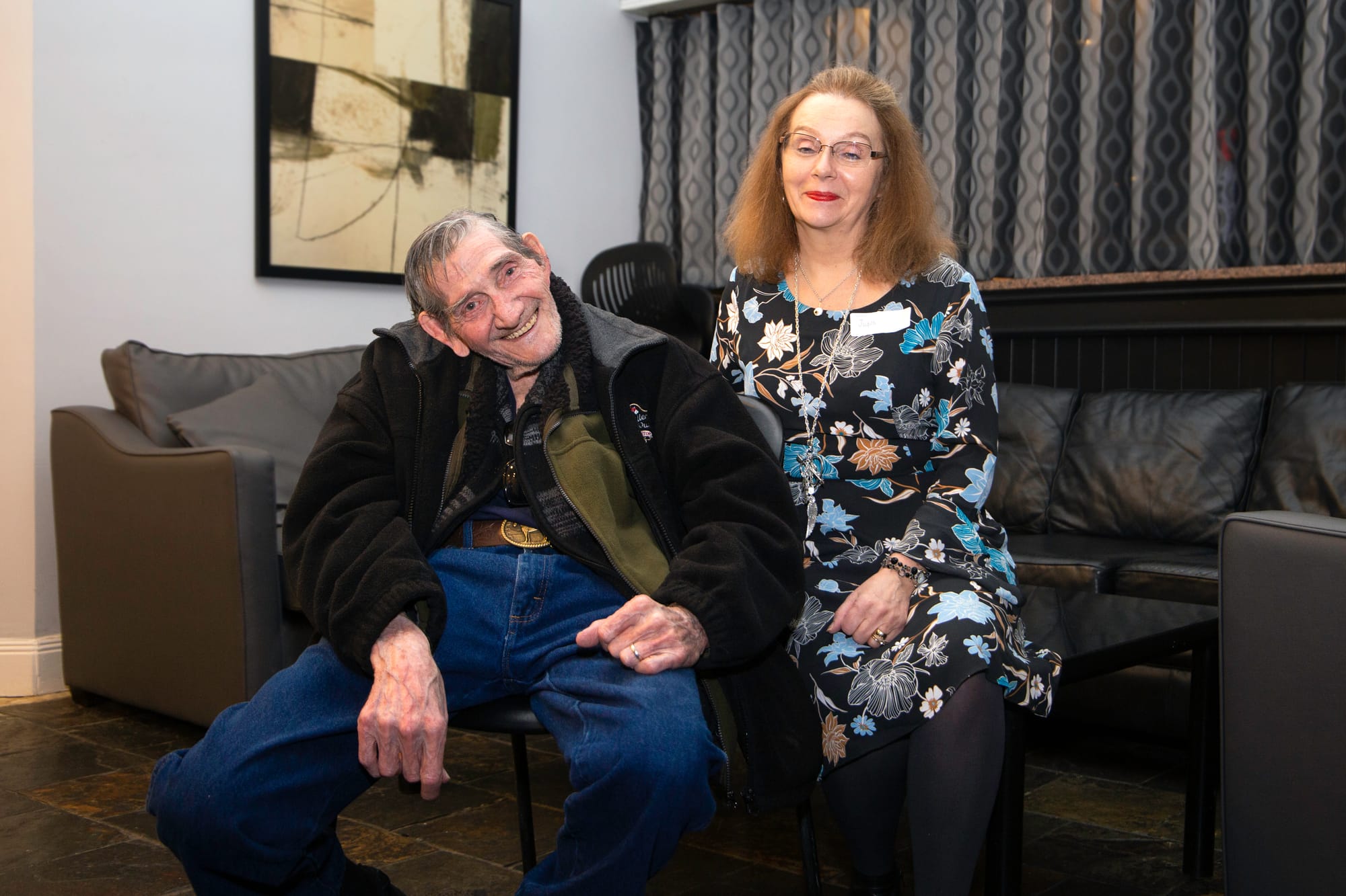 An elderly man sits smiling happily at the camera with a middle aged woman perched on the table next to him, also smiling.