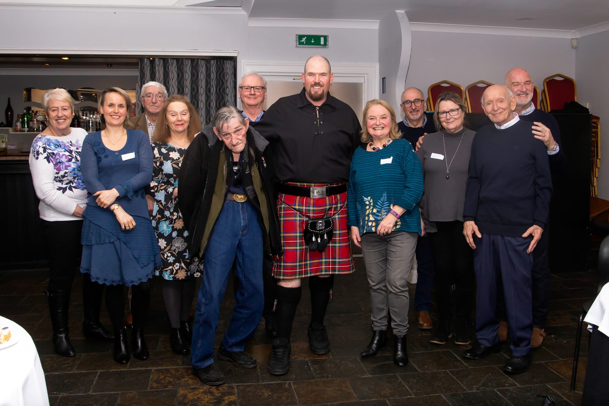 A grup of people stand smiling at the camera with a tall kilted man in the centre next to his elderly father.