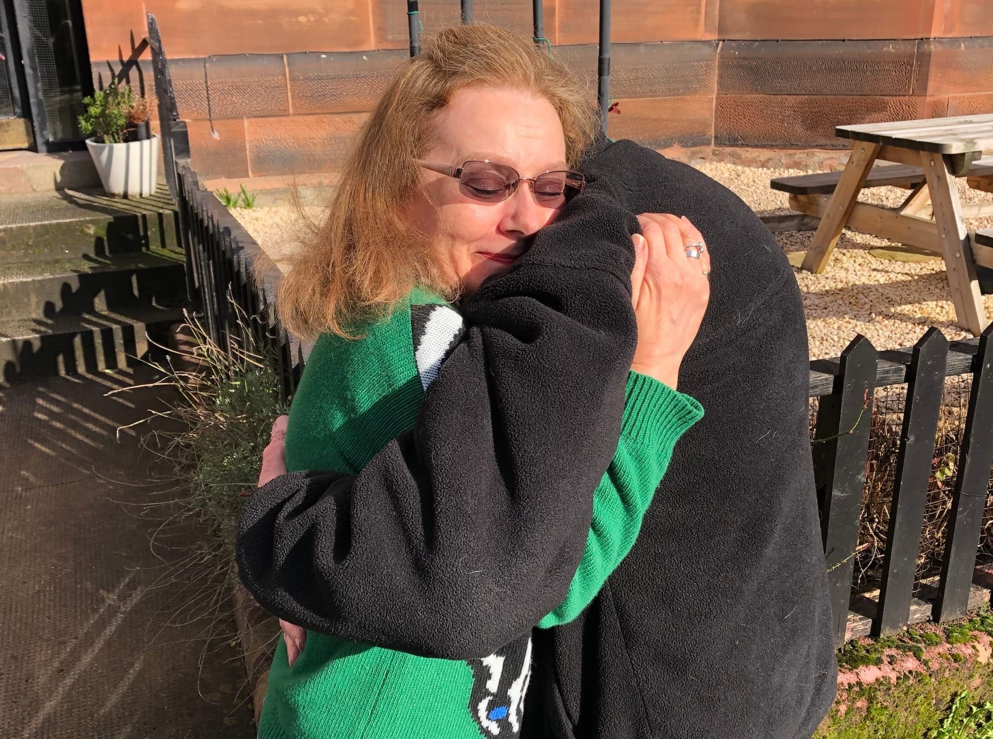 A woman with a green jumper, red hair and glasses hugs and elderly man in a black fleece.