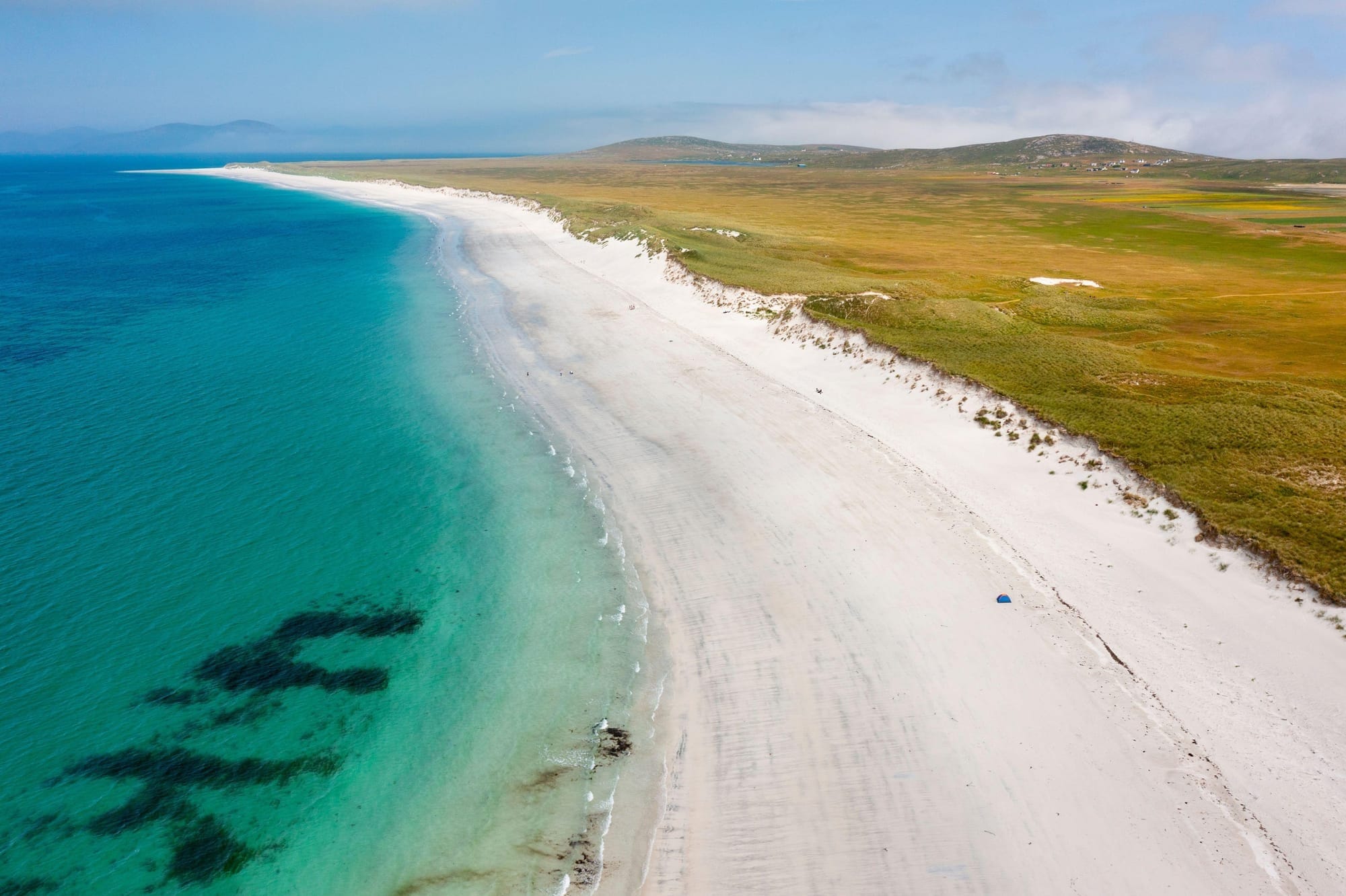 Blue green seas are divided from a flat stretch of greenery by a white sand beach on the Berneray coastline.