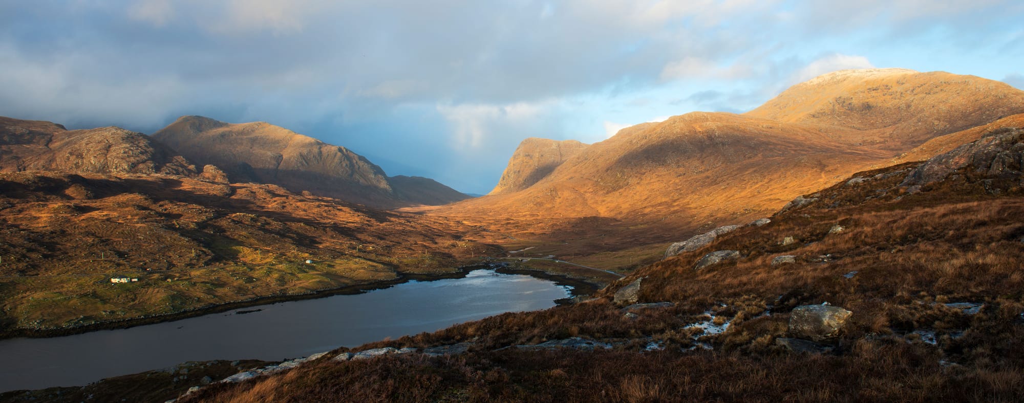 Rugged mountains are lit by the sun with a moody sky behind them. A loch lies below them.