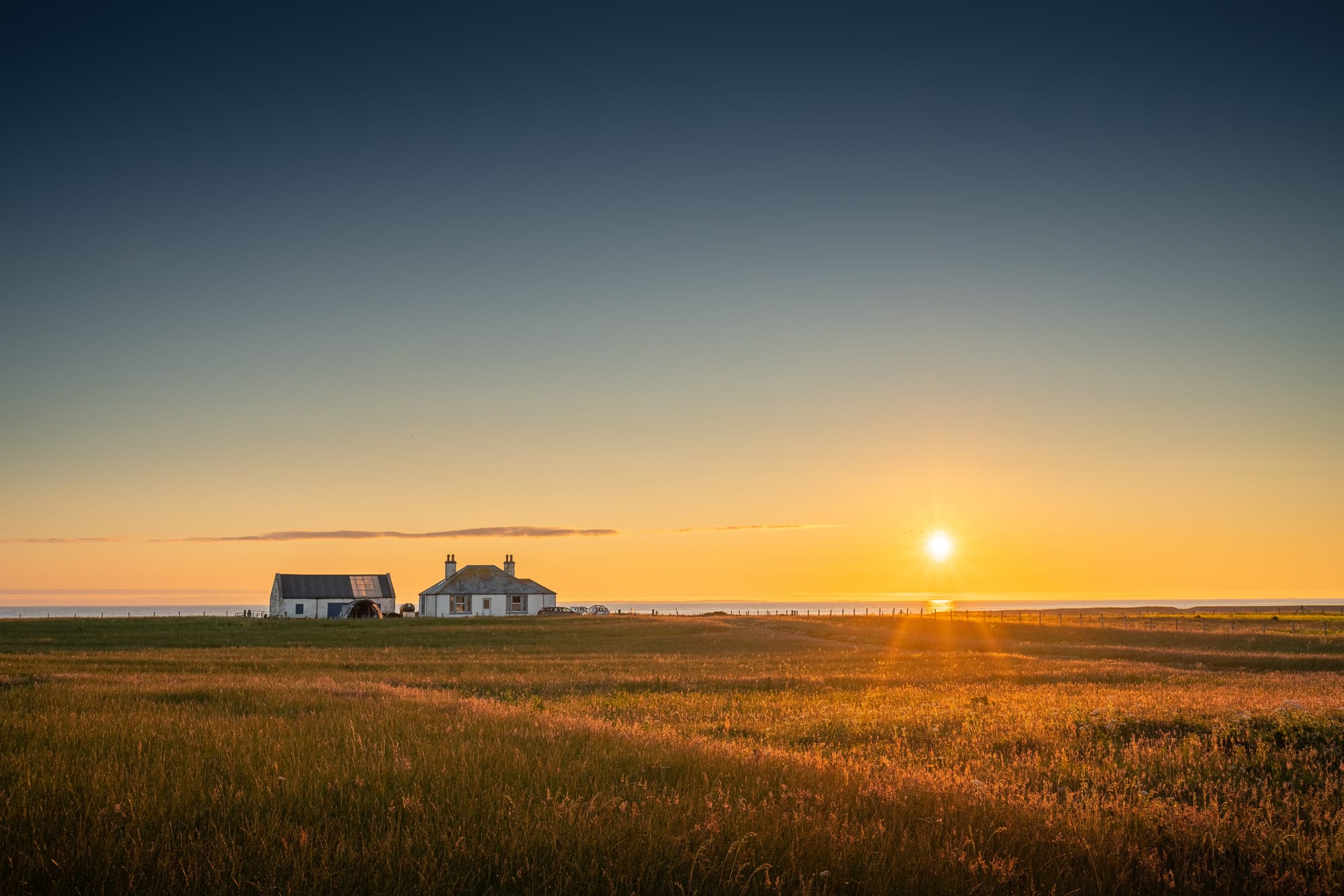 Two small white houses with a field in front of them, lit by a sunset in a clear sky.