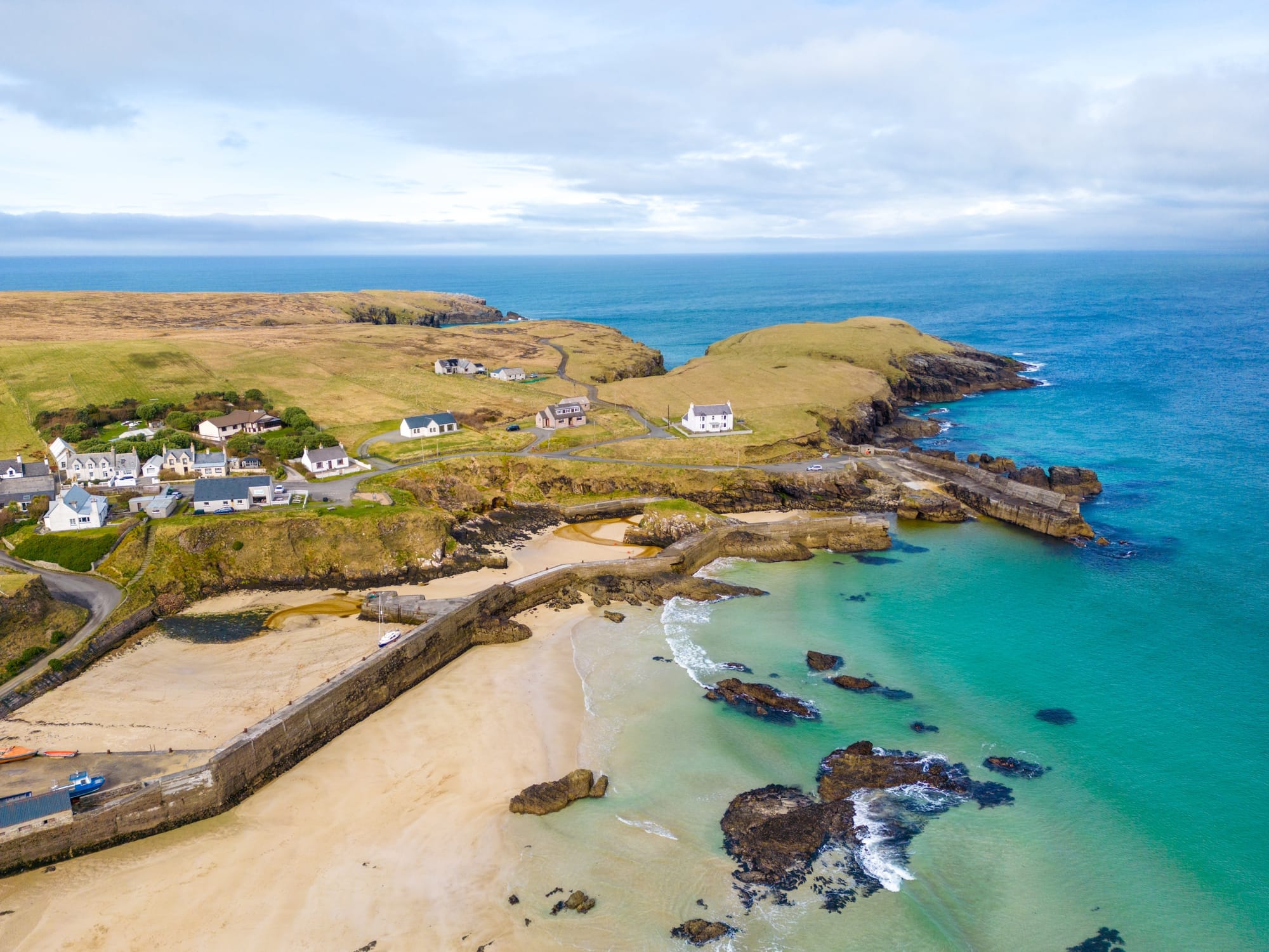 Blue/green seas surrounding the coast and sandy beach below the village of Port of Ness on Lewis. A few small houses sit along the coast above the harbour.