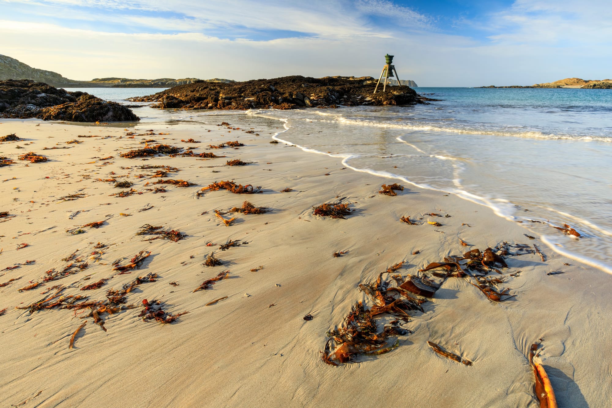 The Time and Tide Bell sits on rocks just off the sandy Bosta Beach on Great Bernera. The beach ad rocks are strewn with seaweed.