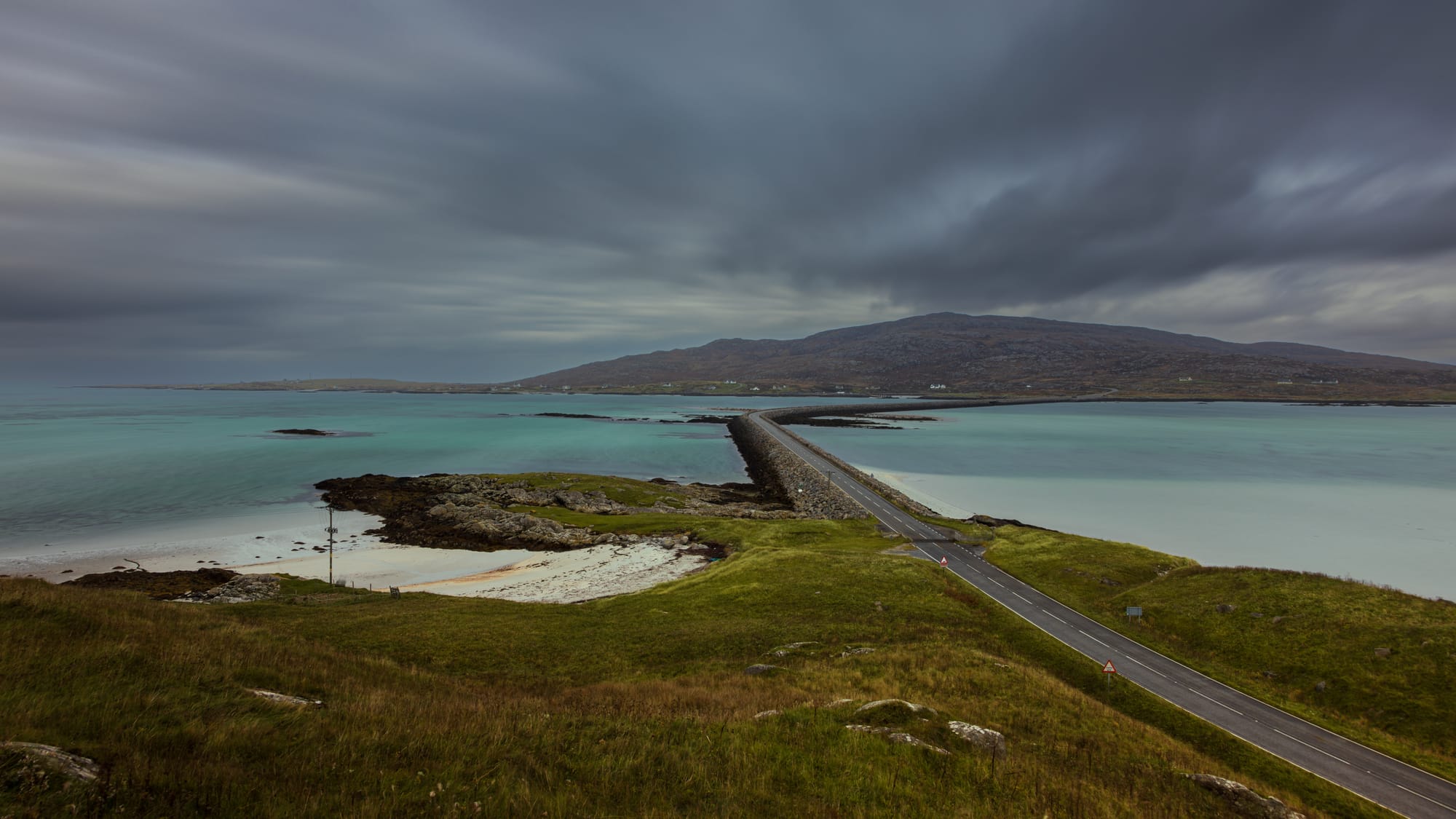 Dark, stormy skies above the long causeway that links Eriskay and South Uist. Flat, blue green seas lies either side of the causeway.