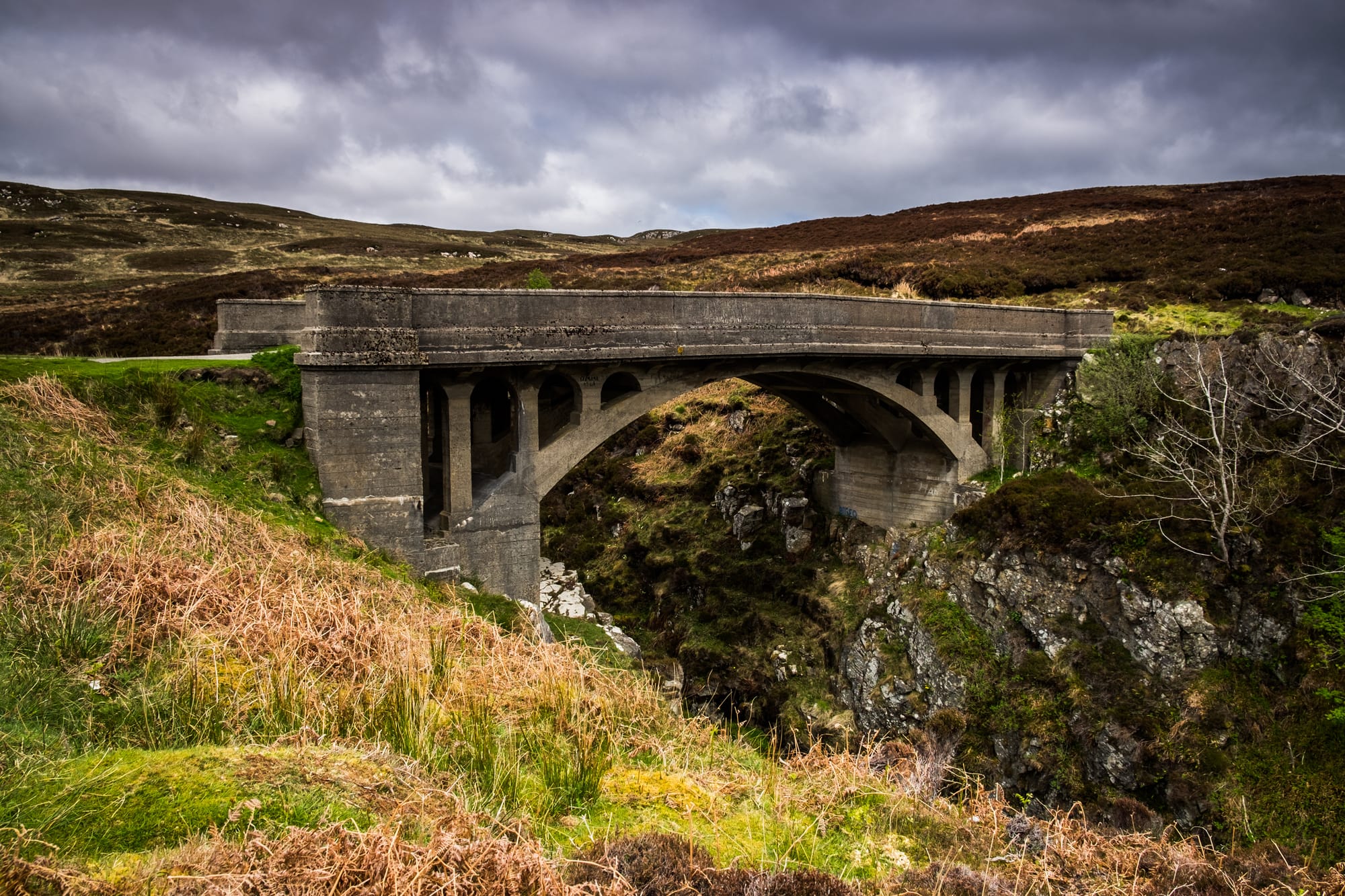 A stone bridge sits above rocky slopes. A road is visible only on one side of the bridge.