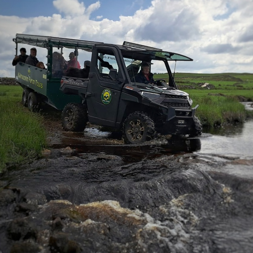 Visitors enjoying a Kilmacolm Farm Tours safari in Renfrewshire, travelling through a shallow stream in an all-terrain vehicle with a covered passenger trailer