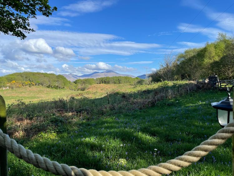 There is a rope fence in the foreground, then grass dotted with wildflowers looking out towards hills in the distance.