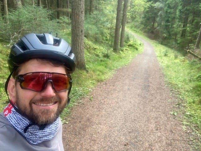 A man in a black cycling helmet and sports sunglasses smiles at the camera on a wooded trail.