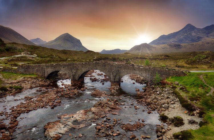 An old stone bridge with three arches over a shallow stony river with the sun setting over mountains in the background.