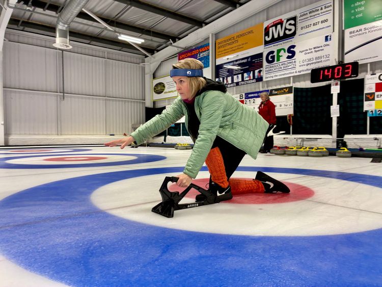 A woman with short blond hair wearing a blue headband and a green jacket is crouched on the ice of a curling rink with arm outstretched after releasing a curling stone.