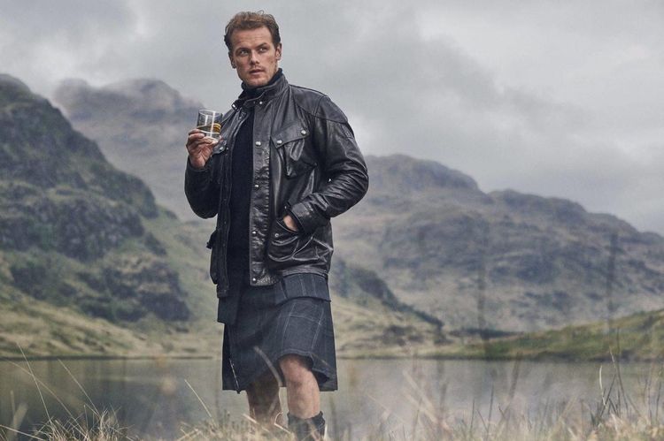 Sam Heughan drinking whisky by a loch. He is wearing a kilt and a leather jacket. 