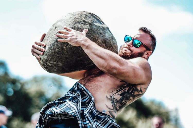 A shirtless man in a kilt lifting a boulder.