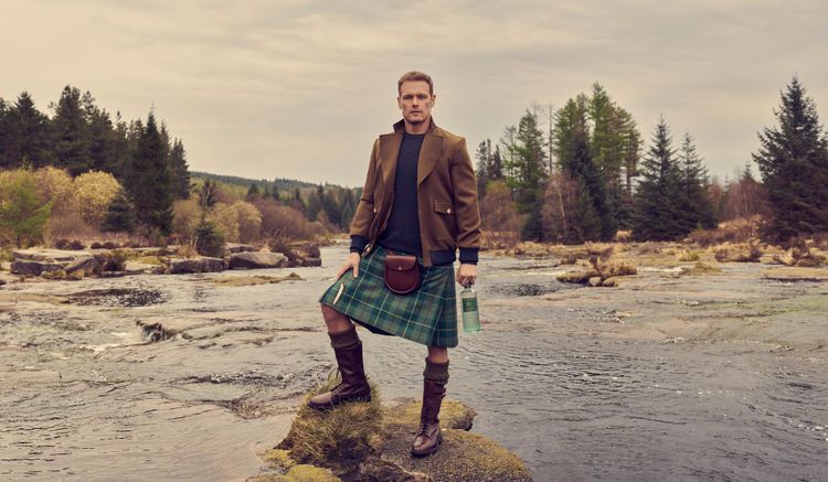 The actor, Sam Heughan, stands on a stone in the middle of a river. He is wearing a kilt with a leather sporran and brown jacket. He holds a bottle of his gin, The Sassenach.