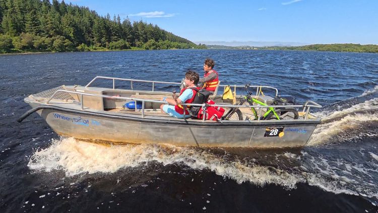 Chris and his friend crossing a loch in a boat. It's a bright day and there are green trees in the background.