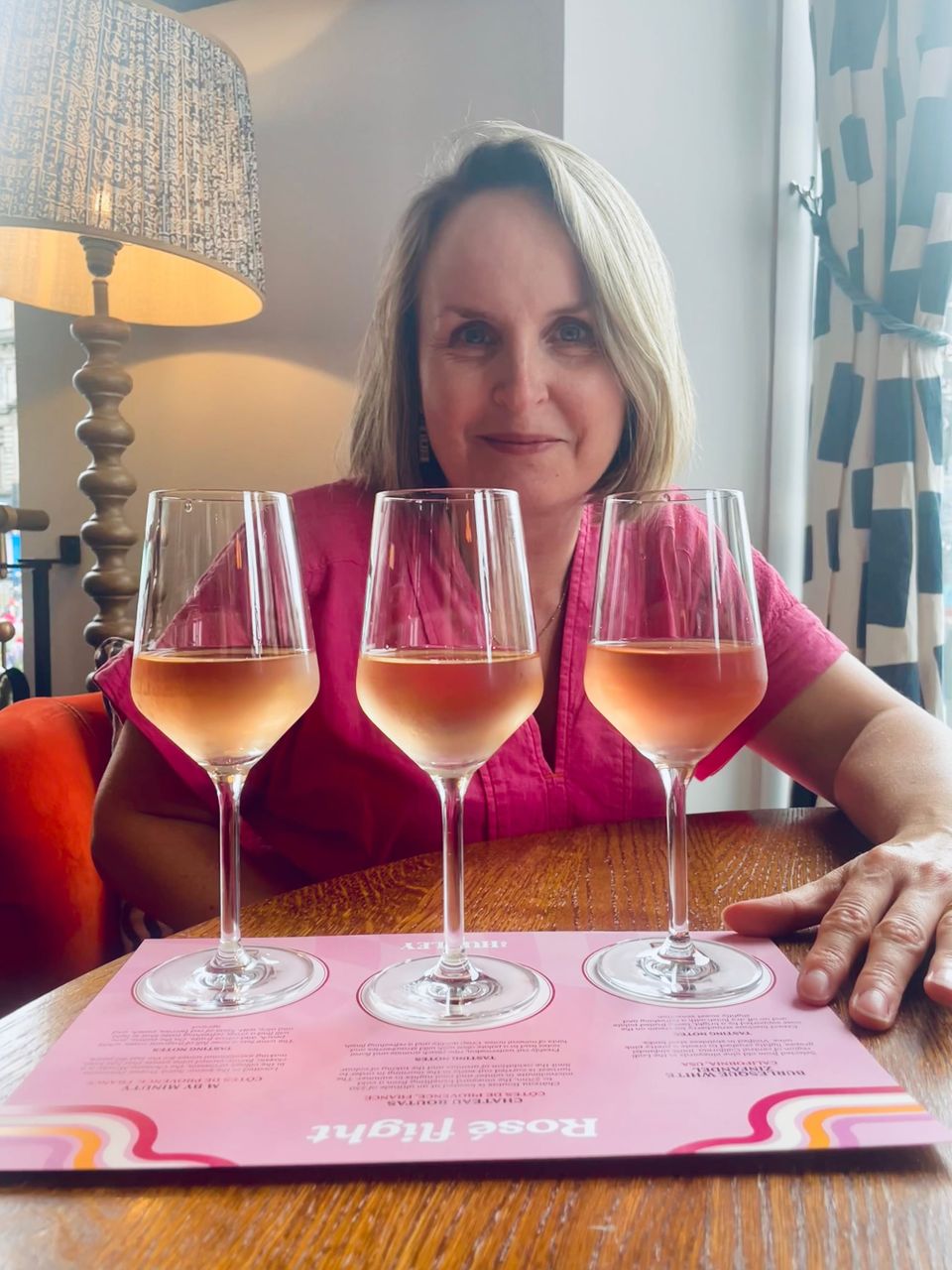 A flight of three different shades of rosé on pink brochure on a wooden table with a blond woman in a pink dress smiling behind them.
