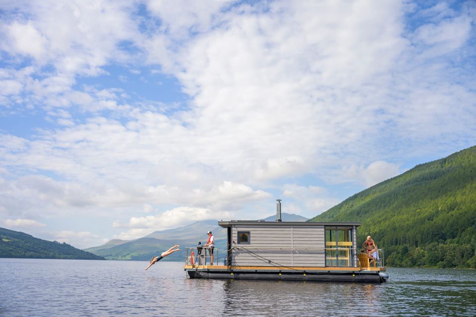 Person diving into Loch Tay from a floating sauna. The loch is surrounded by green hills and there is a cloudy blue sky.