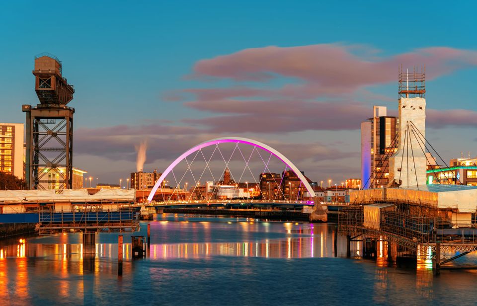 The river Clyde flows into Glasgow beneath a purple lit arching bridge with remnants of the shipbuilding past on either side of the river as well as modern flats.