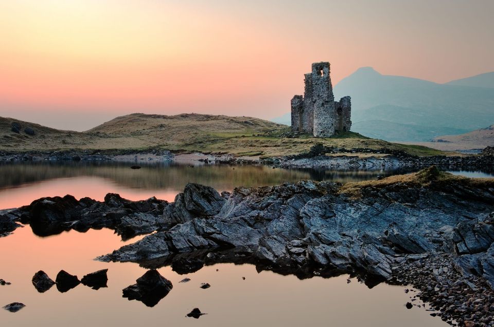 The ruined Ardvreck Castle. In the foreground there are rocks and a loch, and the sky is pink and yellow.