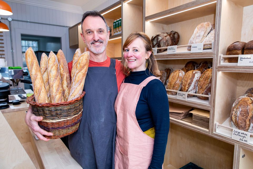 The owners of Wild Hearth Bakery stand behind the counter holding a basket of baguettes. 