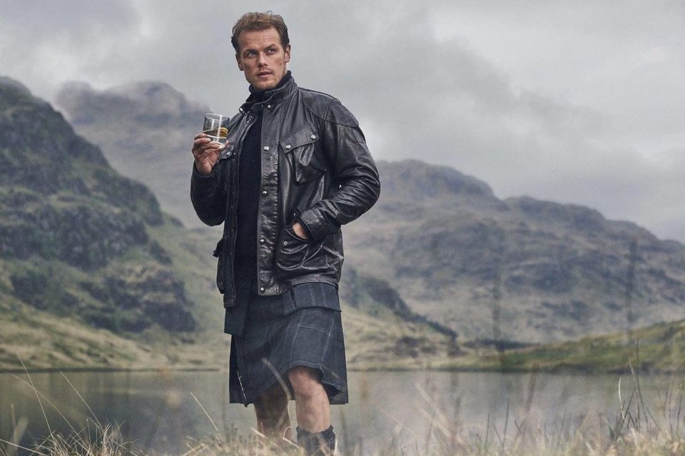 Sam Heughan drinking whisky by a loch. He is wearing a kilt and a leather jacket. 