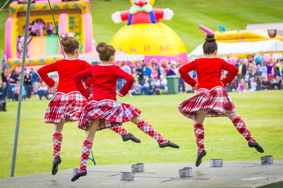 Three girls wearing red jackets and kilts, highland dancing on a stage.