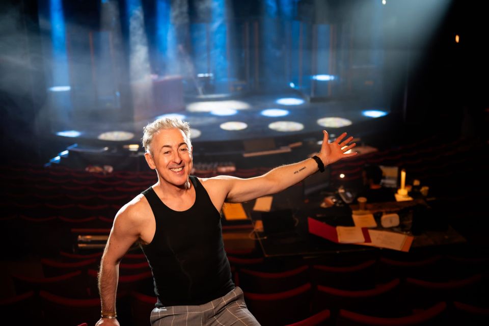 Alan Cumming smiles, gesturing to an auditorium and stage behind him.