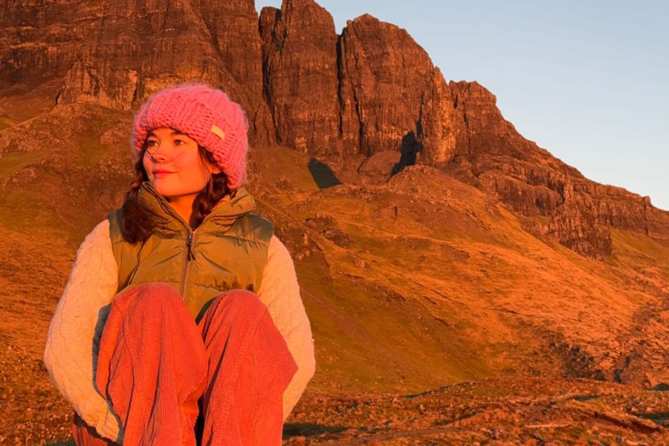 Niamh sits in warm clothes with a rocky landscape behind her at sunset.