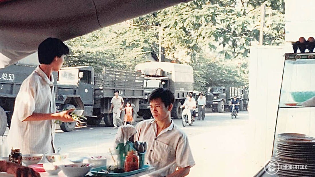 I had breakfast with the translator at a roadside food stall on Sunday morning, April 30, 1995. There are military trucks and personnel in the background, reflecting the blend of daily life and the significant national celebrations about to take place. Street food stalls like this were (and still are) a staple of Vietnamese urban life, offering affordable meals such as phở, bún bò, or bánh mì.