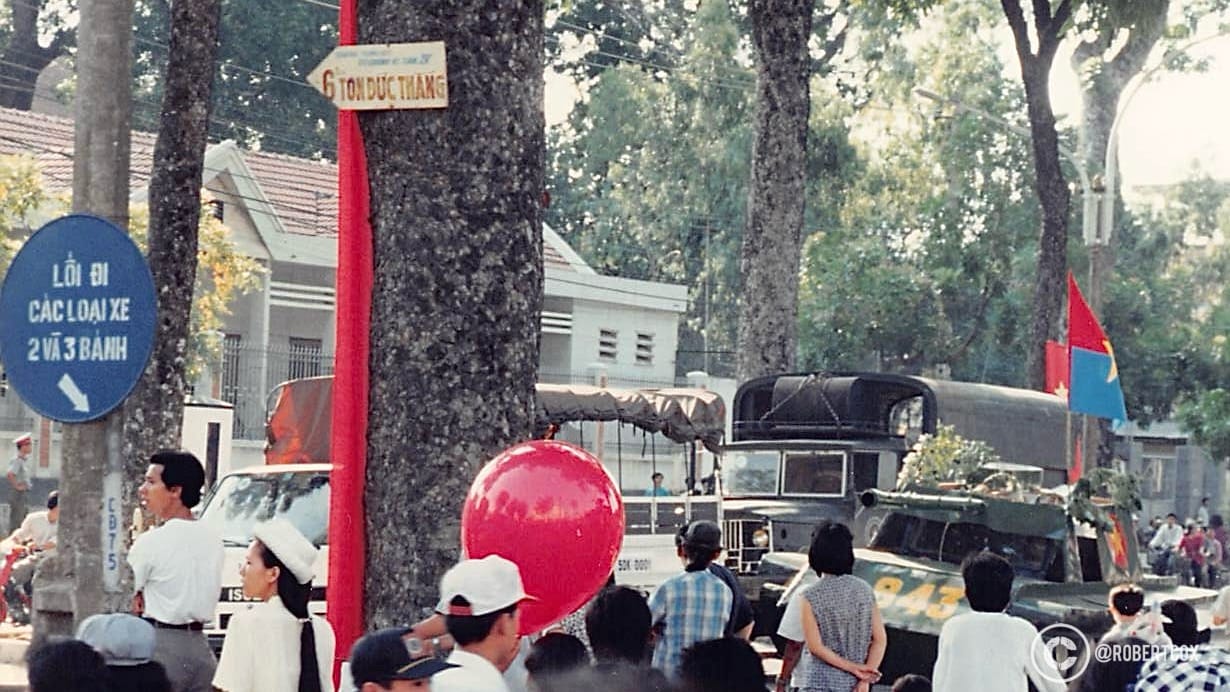 A scene from the parade celebrating the 20th anniversary of Vietnam’s reunification (April 30, 1975 - April 30, 1995). A military vehicle, is part of the parade procession, adorned with greenery and Vietnamese flags, symbolizing national pride. The street signs indicate directions: one sign reads “Lối đi các loại xe 2 và 3 bánh,” which translates to “Path for 2 and 3-wheeled vehicles,” pointing to the right, and another sign above it reads “6 Tôn Đức Thắng,” indicating a street name and direction to the left. Tôn Đức Thắng is a notable street in Ho Chi Minh City, named after a former president of Vietnam.