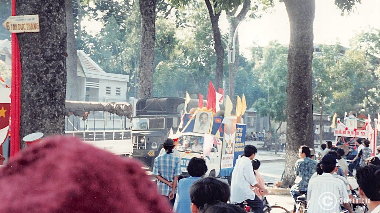 A military vehicle, adorned with Vietnamese flags and other colorful banners, moving through the parade route. A large portrait of Ho Chi Minh, the revered leader of Vietnam’s independence movement and the country’s first president, is prominently displayed on the vehicle. The street is tree-lined, and a sign reading “6 Tôn Đức Thắng” is visible, indicating the parade staging area is taking place on or near Tôn Đức Thắng Street, a major road in Ho Chi Minh City named after a former Vietnamese president. (April 30, 1995).