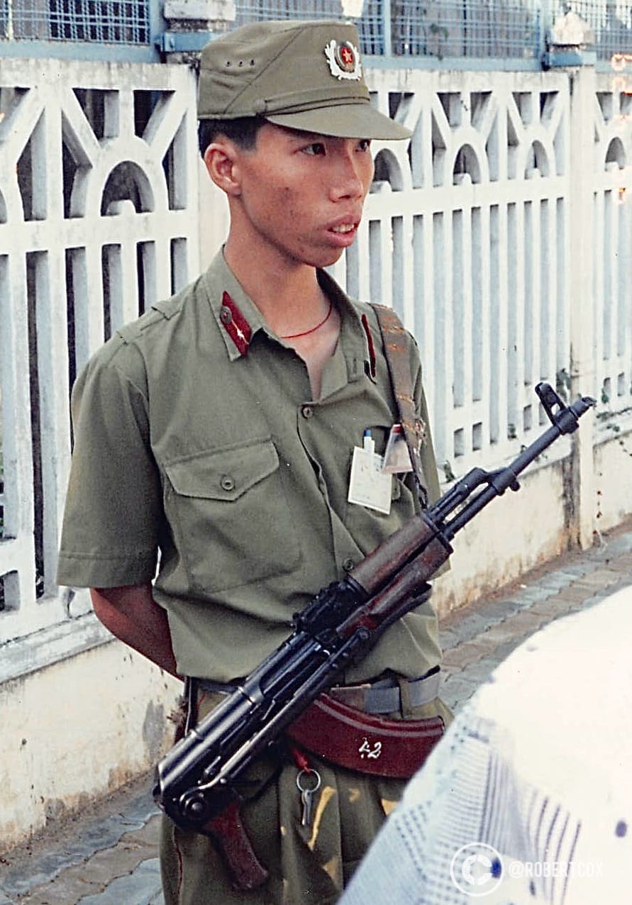 A soldier, holding an AK-47 rifle, a common weapon used by Vietnamese forces during and after the Vietnam War. The uniform includes a cap with a red star and wreath insignia, red epaulettes with stars, and a badge, indicating a rank within the military.