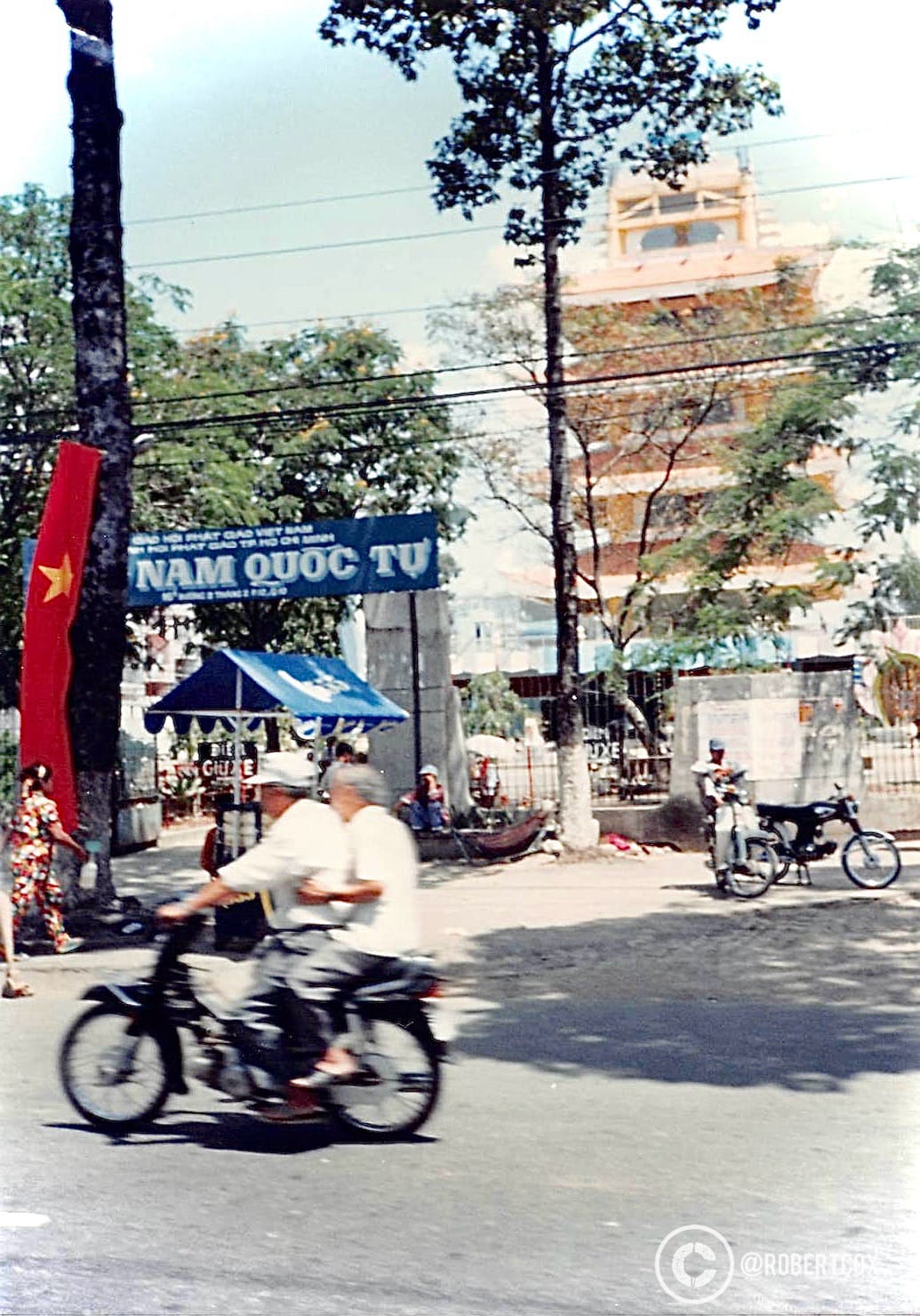 A photo taken from inside a taxi on the way to the airport, looking out through the window, providing a snapshot of daily life in Saigon during your departure, on April 30, 1995. The blue sign says “Ho Chi Minh City Cultural Fair Delegation from April 27 to May 2 / Nam Quốc Tử.” (The location of the fair).