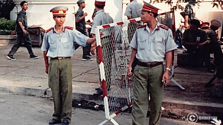 Two members of the Vietnamese police managing the parade to ensure public safety during the event. The uniform features a badge with a star and wreath, typical of Vietnamese law enforcement insignia during that period. They are handling a metal barricade with red and white colors, possibly setting it up or adjusting it to manage the crowd or direct the flow of the parade.