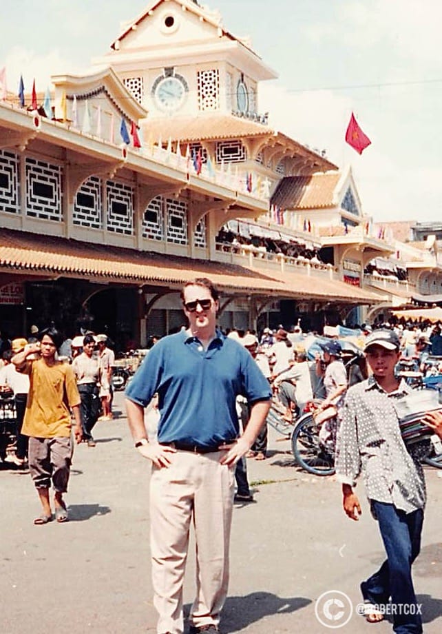 Me, standing in front of the Bến Thành Market, a historic and iconic landmark in Ho Chi Minh City, during the celebrations for the 20th anniversary of Vietnam’s reunification. The market is recognizable by its distinctive architecture, featuring a large clock tower at the main entrance, traditional Vietnamese design elements like the curved roof, and the intricate latticework on the upper facade. Built in 1912 by French colonial authorities, the market is one of the city’s oldest and most famous, known for its bustling atmosphere and as a central hub for locals and tourists to buy everything from fresh produce to souvenirs. (April 29, 1995)