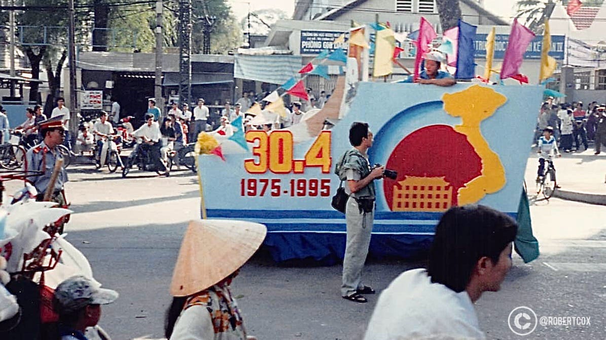 A float entering the parade route prominently displays “30.4 1975-1995,” marking the reunification anniversary, and features a large outline of Vietnam’s map in yellow against a red background, symbolizing national pride. The map outline includes a depiction of the Ho Chi Minh Mausoleum, a significant landmark in Hanoi, further emphasizing the patriotic theme. The crowd includes participants on bicycles, some wearing traditional conical hats (nón lá), and others in uniforms, part of the parade’s organizing or security team. (April 30, 1995)