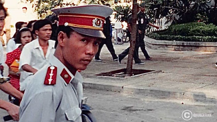 The person in the foreground is a member of the Vietnamese police managing the parade to ensure public safety during the event. The uniform features a badge with a star and wreath, typical of Vietnamese law enforcement insignia during that period.