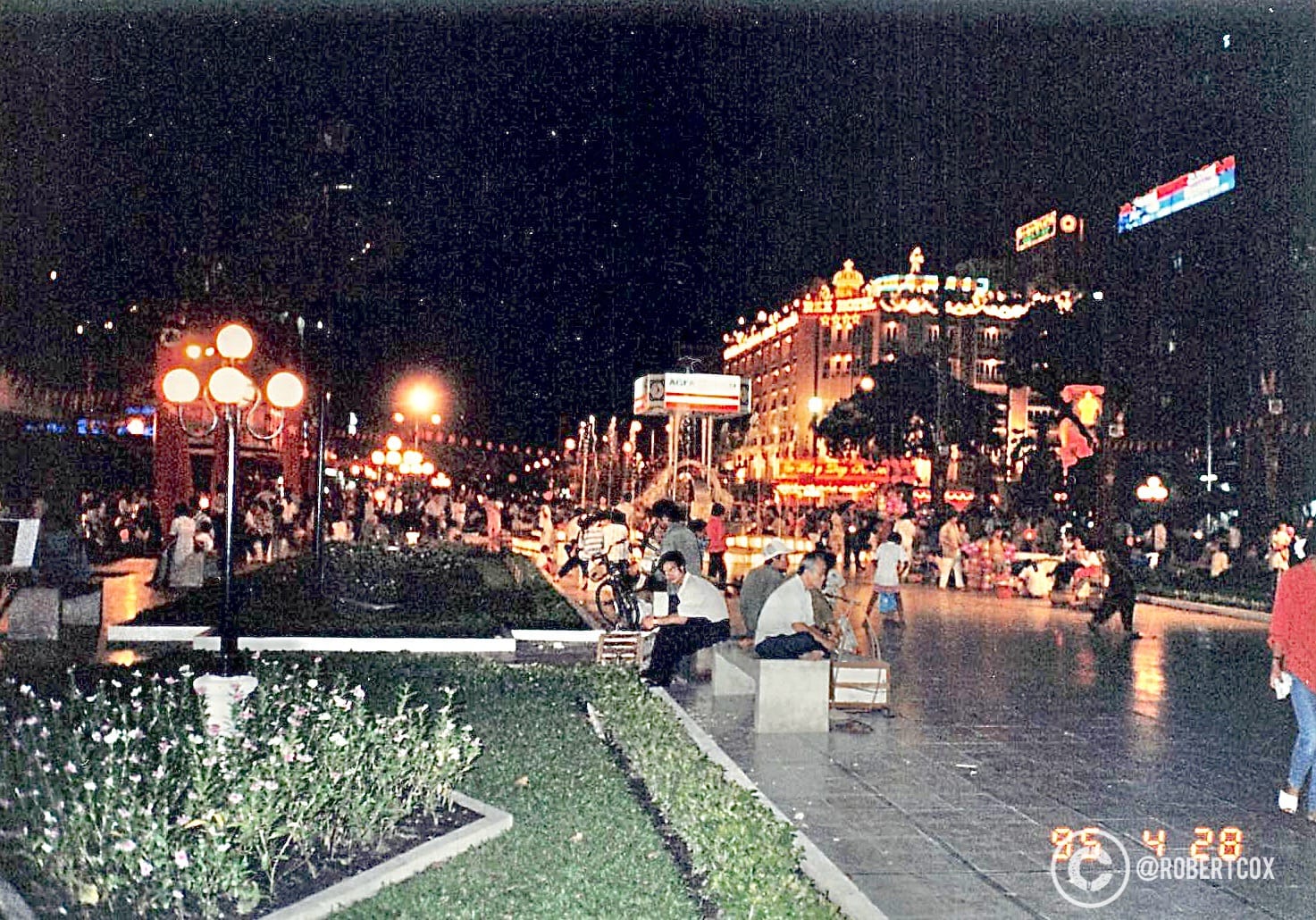 A nighttime scene along Lê Lợi Street in District 1, Ho Chi Minh City, Vietnam. The brightly lit, ornate building in the background is the Hotel Continental Saigon, a historic luxury hotel built in 1880 during French colonial times. The street is bustling with people, street vendors, and cyclists, reflecting the vibrant atmosphere of this central area near the Saigon Opera House and Nguyễn Huệ Walking Street (April 29, 1995).