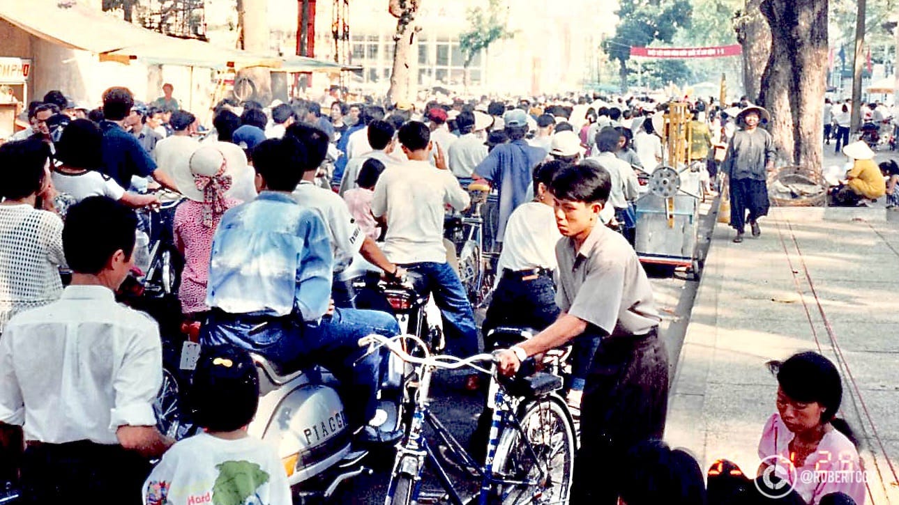 A lively scene from near the parade route in Ho Chi Minh City, Vietnam, on April 30, 1995. A large group of people on motorbikes and bicycles, moving through a tree-lined street adorned with colorful flags, near the parade procession. Some women wearing traditional Vietnamese attire, such as the conical hat (nón lá) and bright dresses.