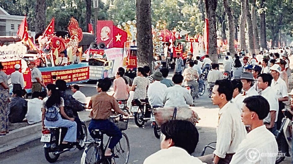 This photo, taken on April 30, 1995, captures a moment in Ho Chi Minh City (Saigon), Vietnam, during the 20th anniversary celebrations of Reunification Day. It shows residents gathered to watch parade groupings as they prepare to enter the official parade route, highlighting the festive atmosphere and the restrictions on public attendance for the main event.