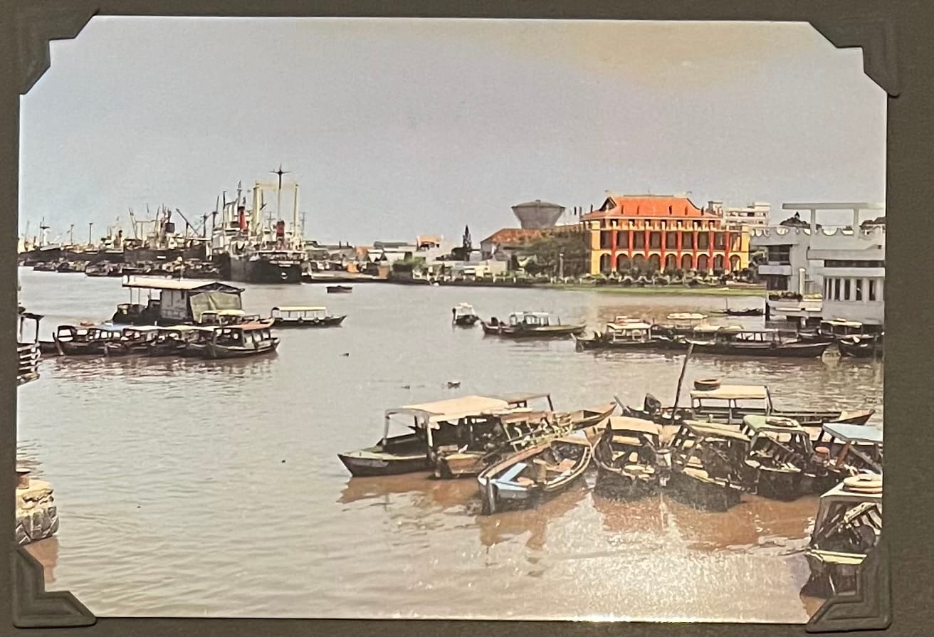 This image is a postcard depicting a scene along the Saigon River in Ho Chi Minh City, Vietnam, with a focus on the riverfront and the bustling activity of boats and ships. The prominent building on the right side of the image is the former Saigon Tax Trade Center (Nhà Thuế Sài Gòn), a French colonial-era structure built in 1887, recognizable by its distinctive red and yellow facade with arched windows and a tiled roof. The building was originally used as a customs house during the French colonial period and later became a commercial center. The Saigon River, a vital waterway for trade and transportation, is filled with numerous boats and ships, ranging from small wooden vessels to larger cargo ships, reflecting the city’s role as a major port. (April 30, 1995)
