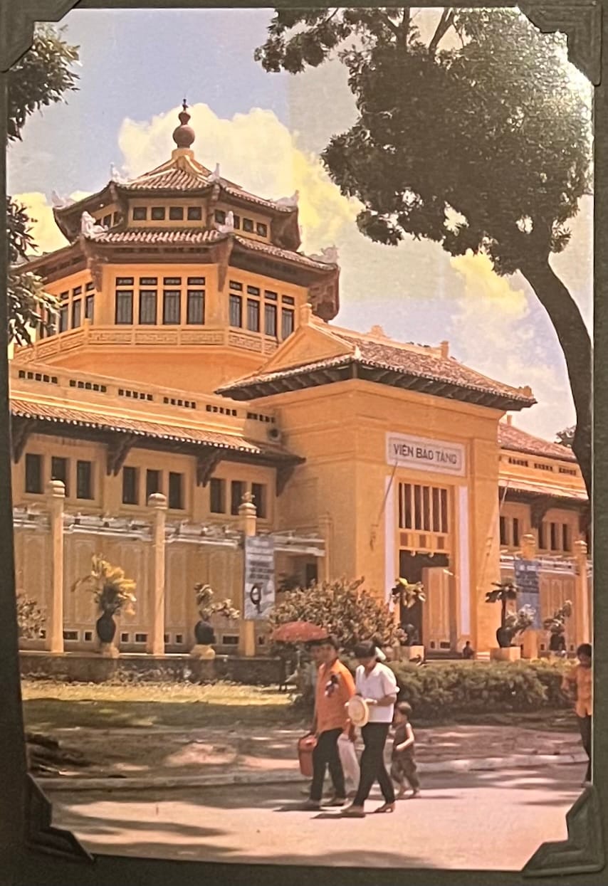 This image is a postcard featuring Vĩnh Nghiêm Pagoda, a prominent Buddhist temple in Ho Chi Minh City, Vietnam. The pagoda is recognizable by its distinctive seven-story tower with a pagoda-style roof, topped with a spire, which is a hallmark of its architecture. The sign “VIỆN BẢO TÀNG” above the entrance translates to “Museum Institute” in Vietnamese, indicating that this section of the pagoda may have been used for cultural or historical exhibitions at the time the image was taken. The image also shows people walking in the foreground, one holding a red umbrella, capturing a casual moment of daily life around the temple. (April 30, 1995)