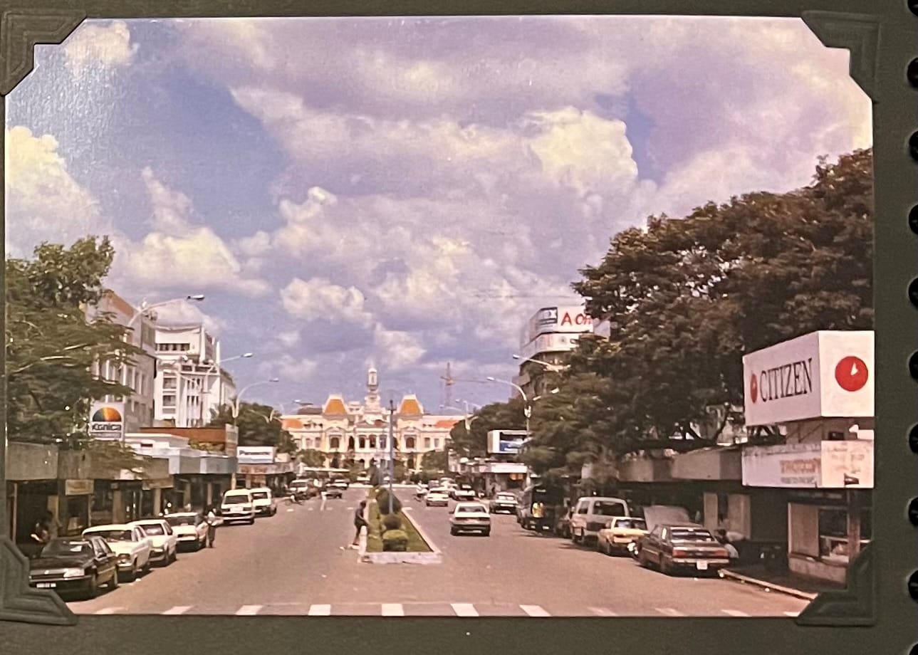 This image is a postcard depicting a street scene in Ho Chi Minh City, Vietnam, with the Ho Chi Minh City Hall (formerly the Saigon City Hall) prominently visible in the background. The City Hall, a French colonial-era building constructed between 1902 and 1908, is recognizable by its distinctive yellow facade, red-tiled roof, and central clock tower with a bell. The street in the foreground is Lê Lợi Boulevard, one of the main thoroughfares in central Saigon, lined with trees, shops, and vehicles, reflecting the bustling urban life of the city. The image captures a mix of older cars and vans, typical of the early 1990s, along with advertisements for brands like Citizen, indicating a growing commercial presence. (April 30, 1995)