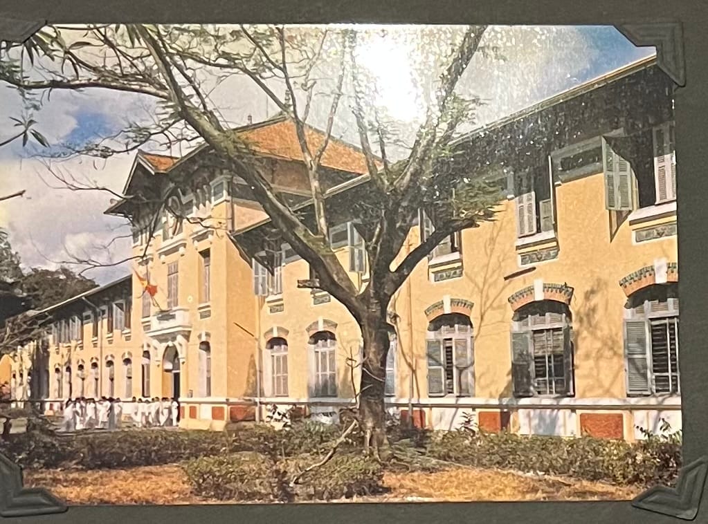 This image is a postcard depicting the Ho Chi Minh City Post Office, also known as the Saigon Central Post Office, in Ho Chi Minh City, Vietnam. The building is a prominent example of French colonial architecture, characterized by its yellow facade, red-tiled roof, arched windows, and ornate details. Designed by French architect Gustave Eiffel, who is also famous for the Eiffel Tower in Paris and completed in 1891, the Post Office is a well-known landmark in central Saigon. The image shows the building’s exterior with a group of people, possibly schoolchildren or a tour group, gathered in front, and a Vietnamese flag flying from the balcony. The surrounding greenery and large tree in the foreground add to the picturesque setting. (April 30, 1995)