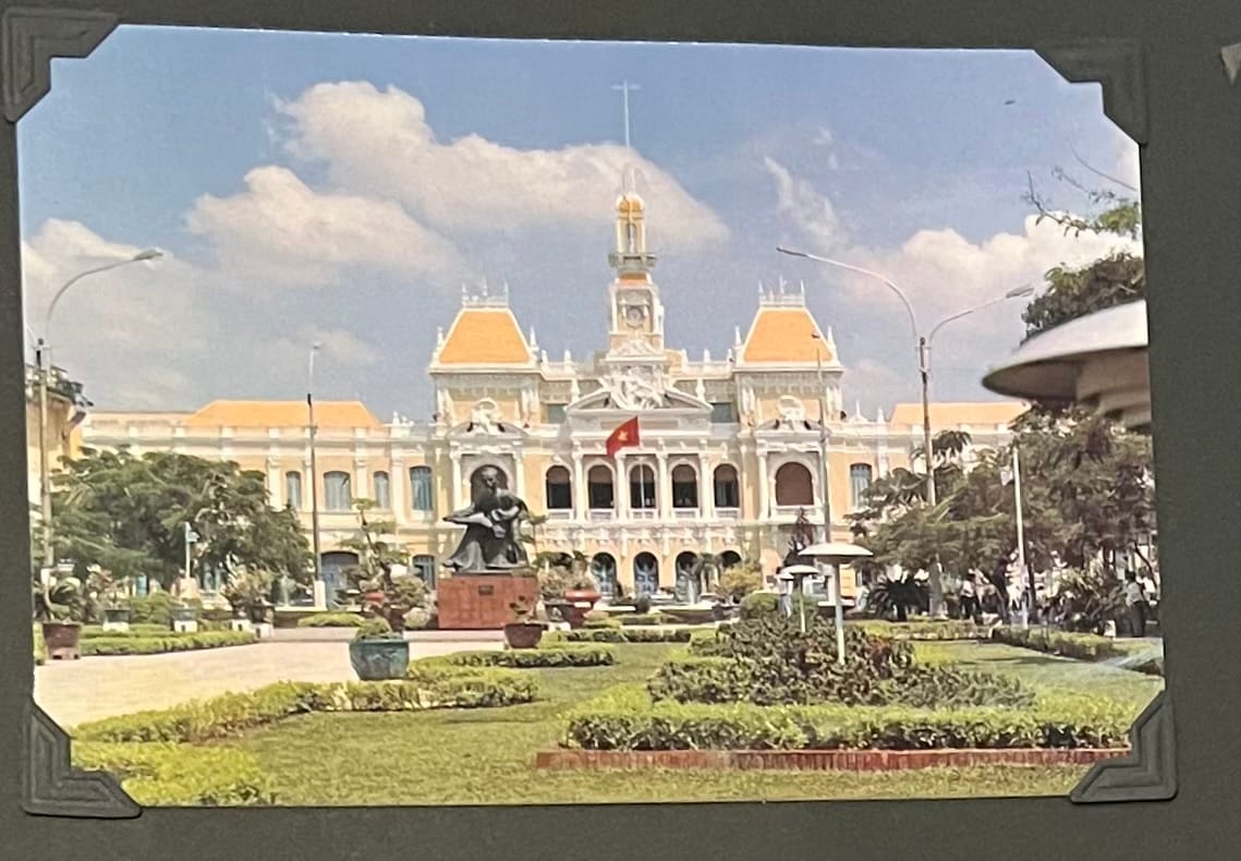 This image is a postcard depicting the Ho Chi Minh City Hall, also known as the Saigon City Hall or the Ho Chi Minh City People’s Committee Headquarters, in Ho Chi Minh City, Vietnam. The building is a prominent example of French colonial architecture, characterized by its yellow facade, red-tiled roof, and central clock tower with a bell. Constructed between 1902 and 1908 during French colonial rule, it originally served as the Hôtel de Ville de Saïgon and has since become a symbol of the city. The foreground features a well-maintained garden with a statue of a seated figure, likely a representation of a Vietnamese scholar or historical figure, surrounded by greenery and pathways. A Vietnamese flag flies above the entrance, reflecting the building’s role under the communist government in 1995. (April 30, 1995)