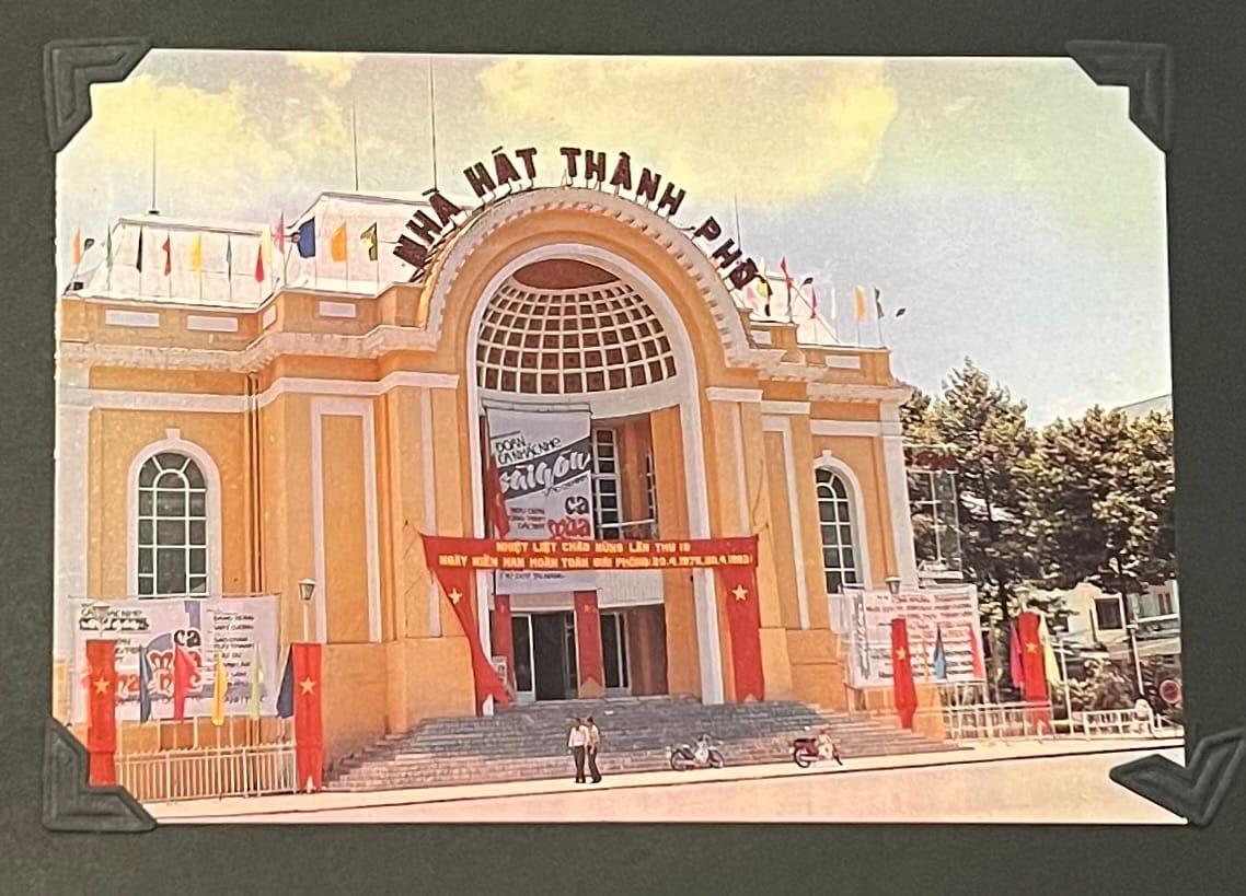 This image is a postcard depicting the Ben Thanh Market (Chợ Bến Thành) in Ho Chi Minh City, Vietnam. The market is recognizable by its distinctive arched entrance and the sign “CHỢ BẾN THÀNH PHỐ” above, which translates to “Ben Thanh Market City.” The building features a yellow facade with a large semi-circular window over the entrance, a design that blends French colonial influences with local architectural elements. The market is decorated with banners, flags, and red streamers, indicating a special event or celebration. (April 30, 1995)