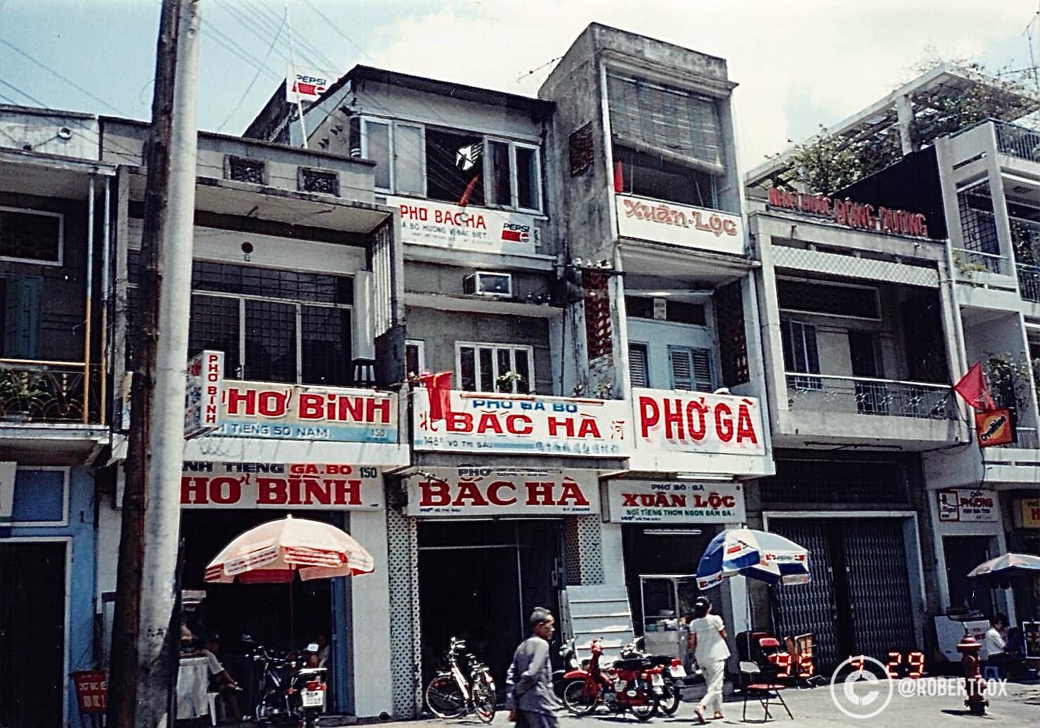 This photo, taken on April 30, 1995, from a taxi on my way to the airport in Ho Chi Minh City, captures a street scene featuring small eateries like “Phở Bắc Hà” and “Hộ Bình.” The image reflects the everyday life of Saigon, with motorbikes, street-side stalls, and locals going about their day, while small Vietnamese flags hint at the ongoing 20th anniversary celebrations of Reunification Day. The Pepsi logo underscores the early influence of Western brands in Vietnam during its economic opening in the 1990s. This final snapshot encapsulates the vibrant street culture and historical moment you experienced in Saigon before departing for Bangkok.