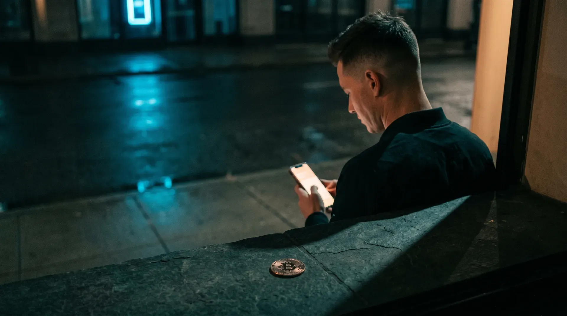 Man in donkere kleding zit 's nachts op stoep, smartphone in handen, Bitcoin-munt op grond