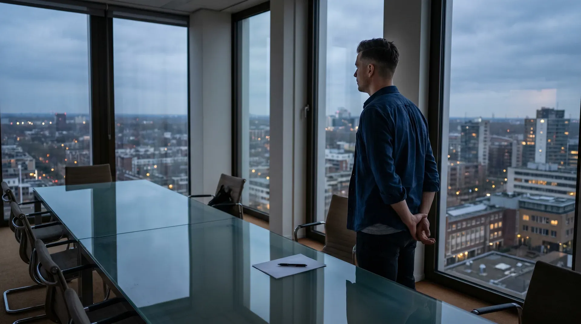 Man in donkerblauw shirt staat bij raam op hoog kantoor, uitzicht over stad met verlichte gebouwen.