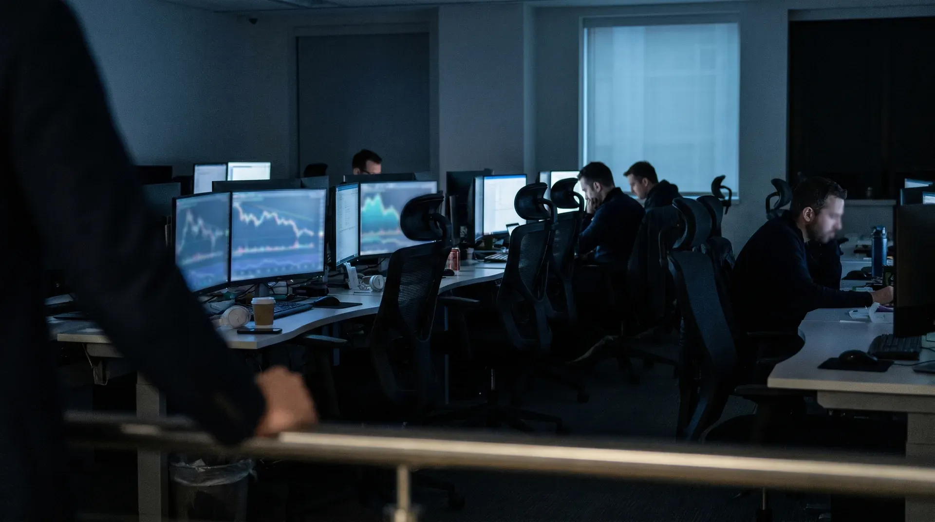 Trading floor met meerdere werkstations, monitoren met grafieken, donkere zakelijke setting met ramen.